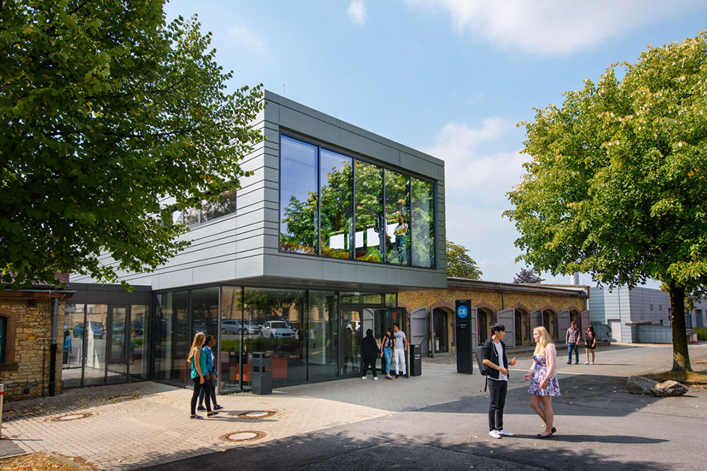 Außenansicht der Caprivi-Lounge ("CaLo") zwischen Bäumen; mehrere Studierende stehen und gehen davor über den Campus.