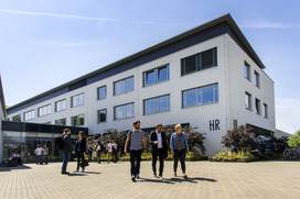 Das Bild zeigt ein dreistöckiges weißes Gebäude mit einem schwarzen Flachdach an einem sonnigen Tag mit blauem Himmel, indem die Bibliothek untergebracht ist. Im Vorderund sind mehrere Studierendengruppen zu sehen, die zum Gebäude hin- oder vom Gebäude weggehen