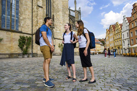 2 Studentinnen und ein Student in sommerlicher Kleidung erkunden den Rathausplatz. Im Hintergrund ist der Dom zu sehen.