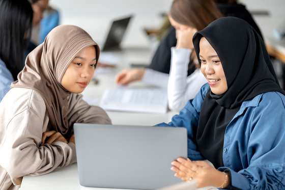 Das Bild zeigt zwei junge Frauen mit Kopftüchern, die gemeinsam an einem Laptop arbeiten. Die Frau auf der linken Seite trägt ein beigefarbenes Kopftuch und einen passenden Pullover, während die Frau auf der rechten Seite ein schwarzes Kopftuch und eine blaue Bluse trägt. Sie lächeln und wirken konzentriert, während sie auf den Bildschirm schauen.  Im Hintergrund sind weitere Studierende in einem modernen Lernumfeld zu sehen. Sie sitzen an Tischen mit Laptops und Schreibunterlagen, was auf eine produktive Arbeitsatmosphäre hindeutet. Die Umgebung ist hell und freundlich, mit natürlichem Licht, das durch große Fenster einfällt.