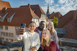 3 fröhlich lächende Studentinnen und 1 Student stehen auf dem Heger Tor und machen ein Selfie. Im Hintergrund sind die Häuser und eine Kirchturmspitze von Osnabrück zu sehen.