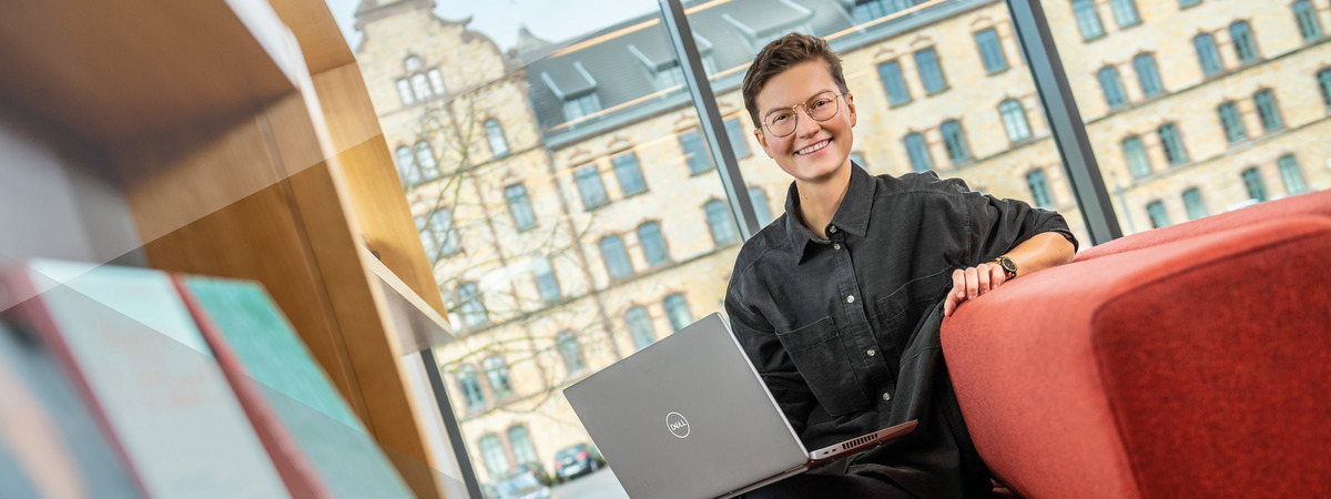 Person sitzt mit Laptop auf einem roten Sofa vor großen Fenstern.