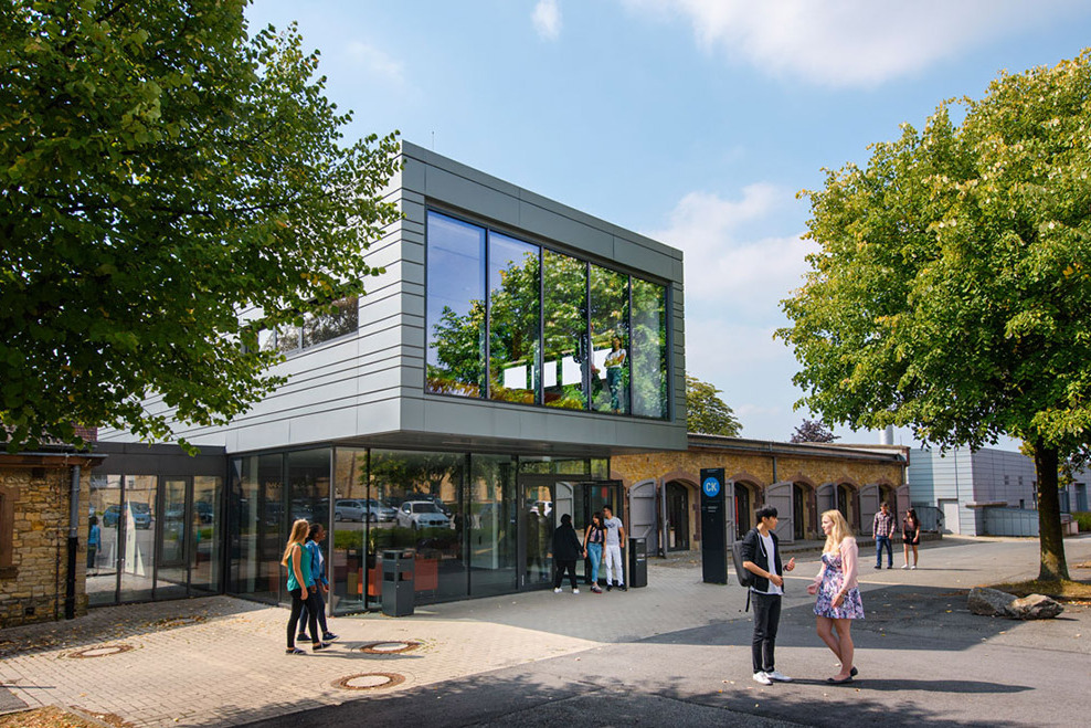Außenansicht der Caprivi-Lounge ("CaLo") zwischen Bäumen; mehrere Studierende stehen und gehen davor über den Campus.